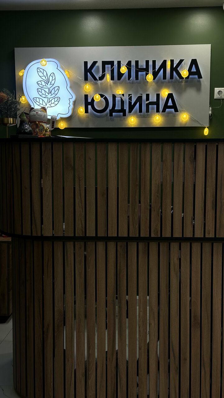 Ресепшен Клиники Юдина — подсвеченная вывеска КЛИНИКА ЮДИНА на тёмно-зелёной стене